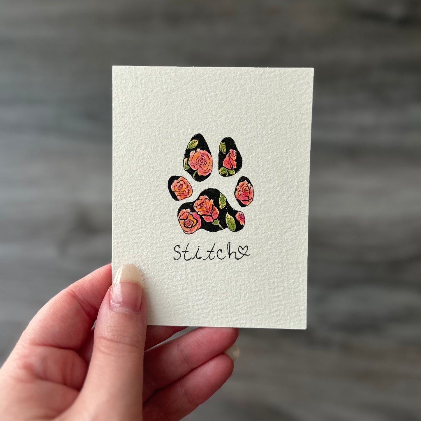 Hand holding a card with a floral paw print design and 'Stitch' text on a gray background