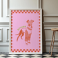 Framed artwork of a stylized line art dog illustration on a pink background and checkered boarder with 'Petunia' text, leaning against a wall.