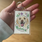 Hand holding a keychain with a dog illustration and floral wreath on a wooden surface