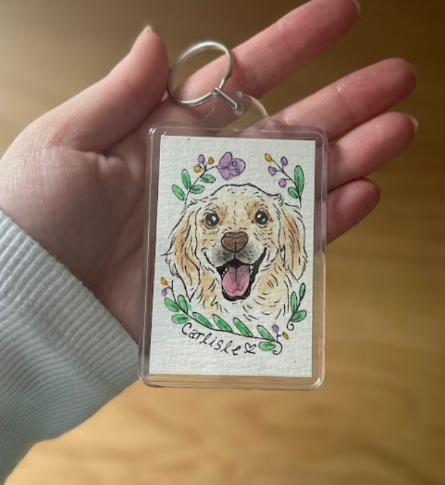 Hand holding a keychain with a dog illustration and floral wreath on a wooden surface