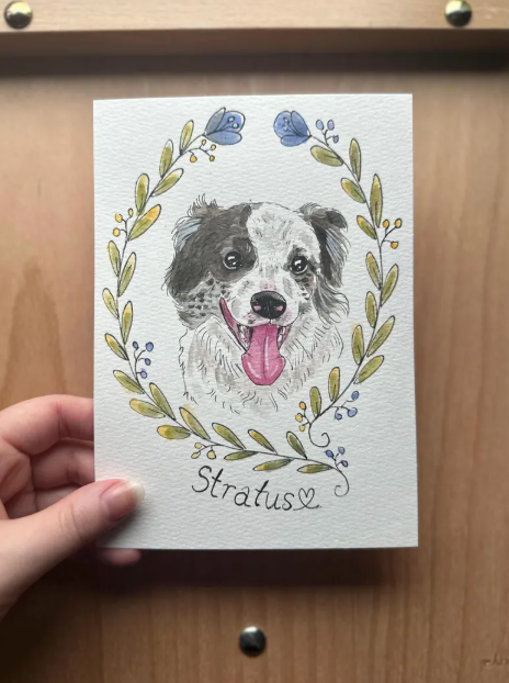 portrait of a watercolor dog illustration and floral wreath held by a hand on a wooden surface