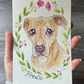 A hand holding a small watercolor-style pet portrait card featuring a tan dog surrounded by green leaves and pink flowers, with the name 'Fred' written below.