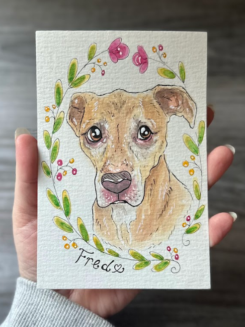 A hand holding a small watercolor-style pet portrait card featuring a tan dog surrounded by green leaves and pink flowers, with the name 'Fred' written below.