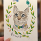 A hand holds a square white art print featuring a watercolor-style cat portrait framed by a green leaf wreath with small colorful flowers. The cat wears a blue and purple bow tie, and the name 'Rupert' is written below in black script.