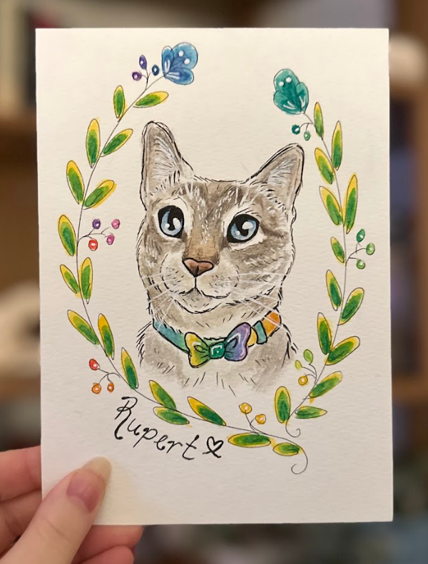 A hand holds a square white art print featuring a watercolor-style cat portrait framed by a green leaf wreath with small colorful flowers. The cat wears a blue and purple bow tie, and the name 'Rupert' is written below in black script.