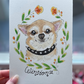 A hand holds a white art print featuring a hand-painted illustration of a chihuahua dog wearing a black collar, surrounded by a green leaf wreath and orange flowers. The name 'Winston' is written in black script at the bottom.