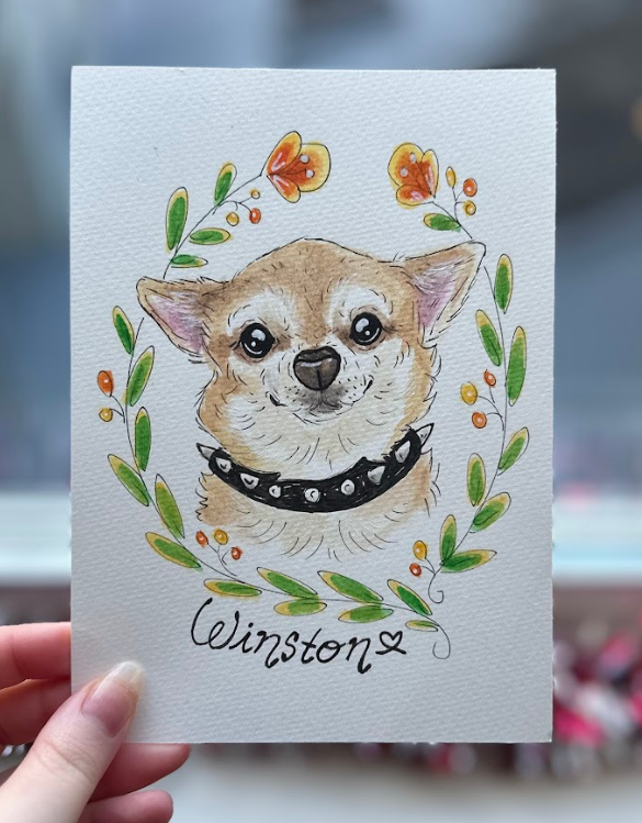 A hand holds a white art print featuring a hand-painted illustration of a chihuahua dog wearing a black collar, surrounded by a green leaf wreath and orange flowers. The name 'Winston' is written in black script at the bottom.