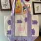 Tote bag with purple cartoon characters and 'Trader Joe's' logo held in front of a wall with framed pictures.