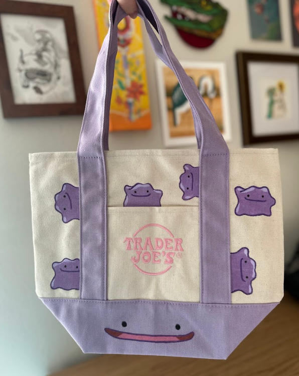 Tote bag with purple cartoon characters and 'Trader Joe's' logo held in front of a wall with framed pictures.