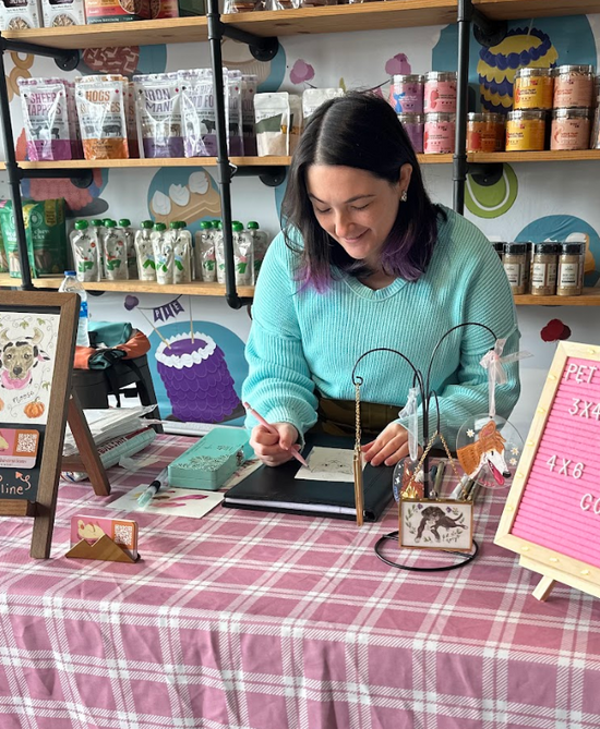Woman in a teal sweater working at an art station with shelves of products in the background.